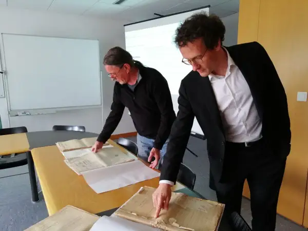 Deux hommes consultent des registres d'état civil aux archives départementales du Morbihan. L'un pointe un document ancien.