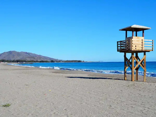 Torre de vigilancia de madera en la playa de Vera, con arena y mar azul. La playa de Vera tiene piedras, pero hoy el sol brilla.