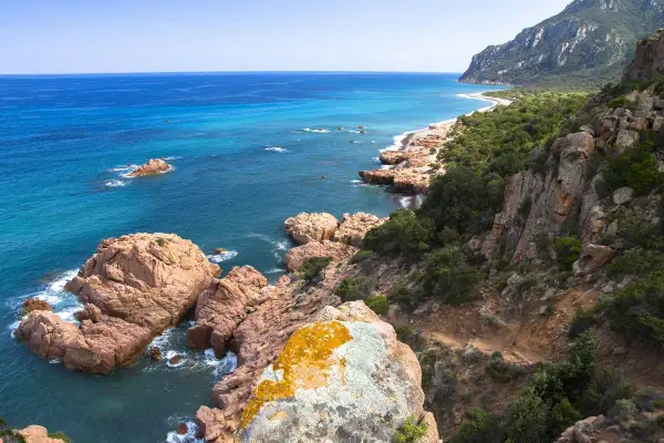 Scogliere rosa e mare turchese sulla costa ovest Sardegna. Un sentiero si snoda tra rocce e macchia mediterranea verso una spiaggia isolata.