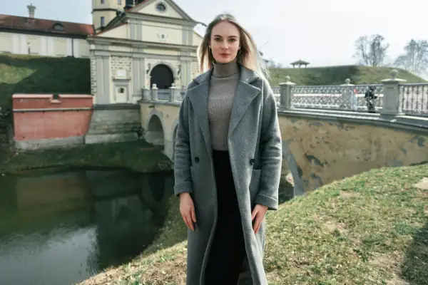 Kobieta w szarym płaszczu i golfie, idealna stylizacja na 14 stopni. W tle zamek i fosą.