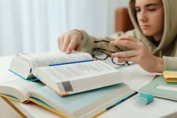 Chłopak w bluzie pochyla się nad książkami, analizując **motywy literackie**. Na stole leżą otwarte tomy, okulary i kolorowe karteczki.