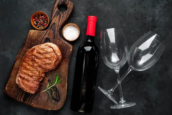 Grillowany stek, butelka wina z regionu Puglia, dwa kieliszki, sól i pieprz na drewnianej desce.
