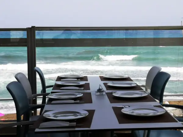 Mesa puesta en el chiringuito la sal, con platos y cubiertos, frente a un mar turquesa y olas rompiendo.