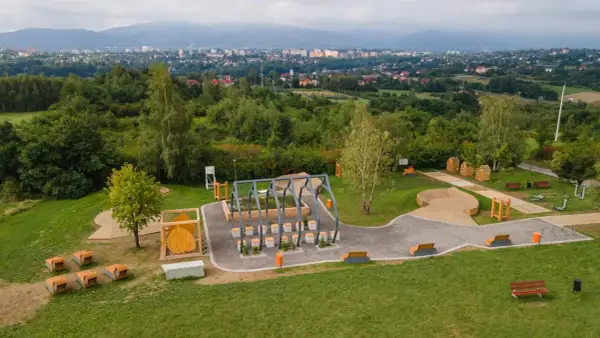 Nowoczesny plac zabaw i siłownia plenerowa, otoczone zielenią, z widokiem na miasto. W oddali widać trzy lipki.