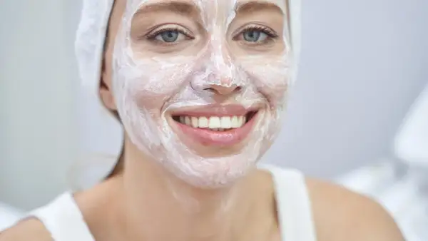 Lächelnde Frau mit weißer Gesichtsmaske und Handtuch auf dem Kopf. Wie oft Maske Gesicht? Regelmäßige Pflege für strahlende Haut.