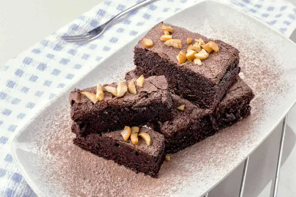 Fette di torta al cioccolato e nocciole, spolverate di cacao e guarnite con nocciole tritate, su un piatto bianco.