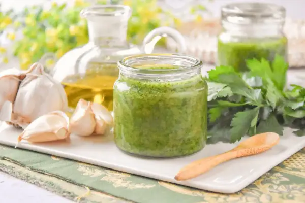 Un frasco de salsa mery verde vibrante, con ajo, aceite de oliva y perejil fresco alrededor.