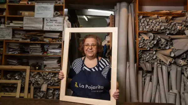 Mujer sonriente en taller, rodeada de rollos de tela y estanterías con materiales. Sostiene un marco de madera en rollo.