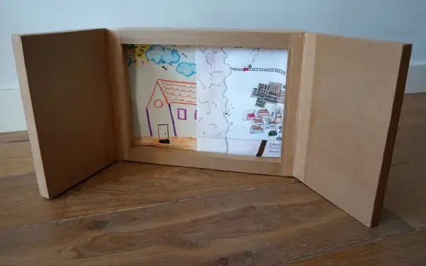 Un kamishibai casero muestra dibujos infantiles: una casa, un paisaje rural y un pueblo con un tren.