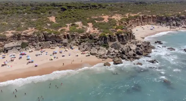 Una cala enebro con acantilados rocosos, arena dorada y aguas turquesas. Gente disfrutando del sol y el mar bajo sombrillas.