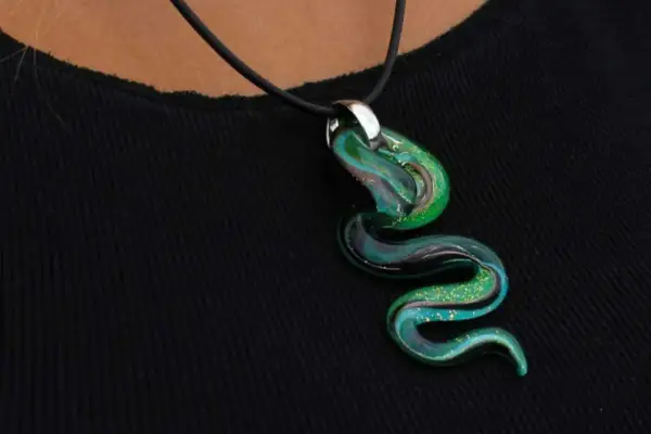 Un serpente sinuoso de **colgante de cristal de Murano** en tonos verdes y azules, con destellos dorados, descansa sobre una tela negra.
