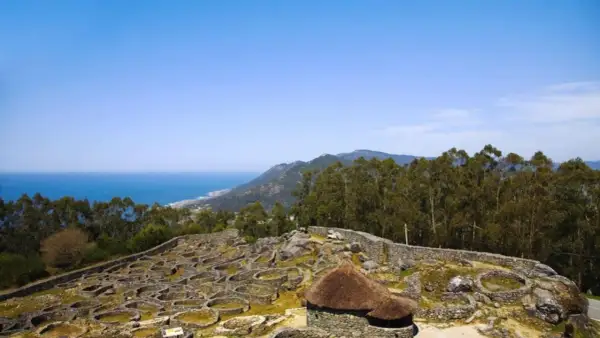 Ruinas circulares de piedra en el monte Santa Tecla, con el mar y montañas al fondo.