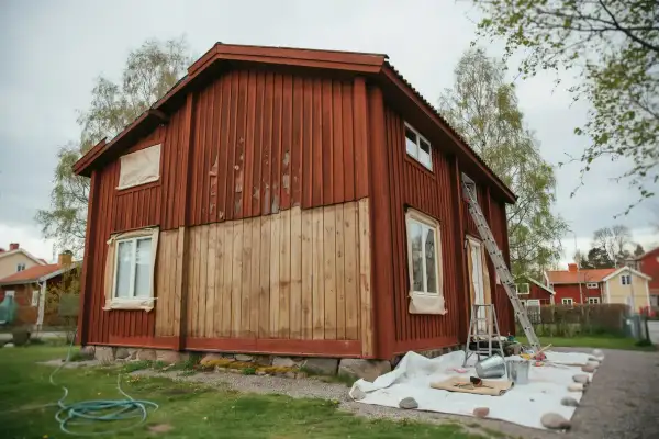 Ett rött trähus målas om. En stege står lutad mot huset, och färgburkar och penslar ligger utspridda på en presenning. Det ser ut som en perfekt dag att måla i solsken.