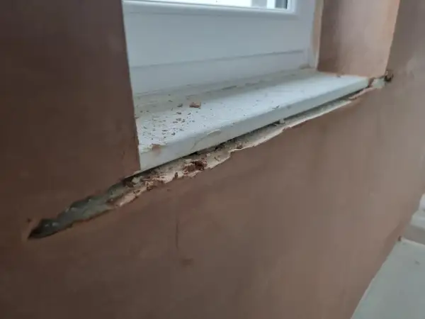 Fensterlaibung verputzen: Rauer Putz umrahmt ein weißes Fensterbrett, das mit kleinen Putzbrocken übersät ist.