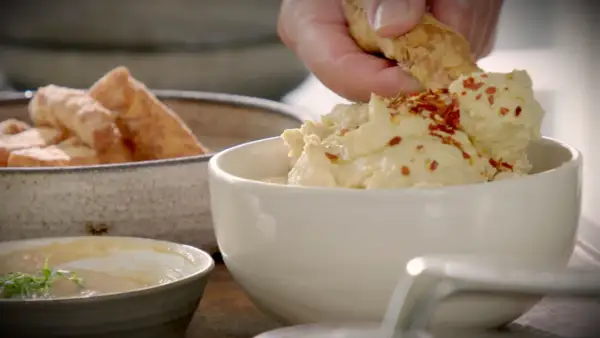 En hand doppar en bit i krämig hummus toppad med chiliflakes. Bredvid står en skål med gyllene halloumi fries.