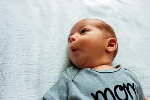 Uroczy niemowlak z kręczem szyi, ubrany w body z napisem "mom", leży na jasnoniebieskim tle.