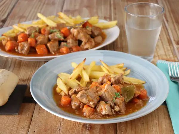 Dos platos de estofado de cerdo con patatas fritas y zanahorias, servidos en una mesa de madera.