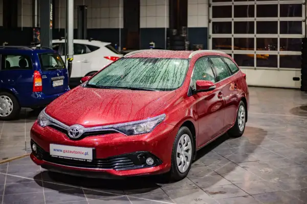 Czerwona Toyota Auris 1.6 benzyna z kroplami deszczu na karoserii, zaparkowana w garażu obok innych samochodów.