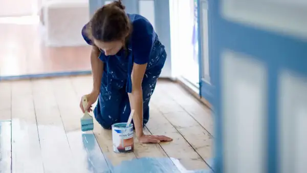 Mujer con mono azul se arrodilla para pintar el suelo de madera de azul claro. Se puede pintar el suelo para renovar la casa.
