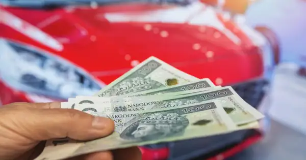Dłoń trzyma banknoty 100 zł na tle czerwonego samochodu. Koszty posiadania auta, w tym podatek od samochodu, mogą być znaczące.