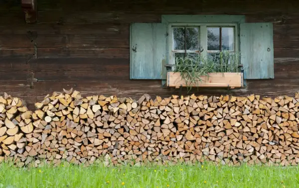 Ein ordentlicher Stapel Holz liegt vor der rustikalen Hauswand. Ein Fenster mit blauen Fensterläden und einem Blumenkasten mit Kräutern darüber.