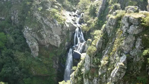 La impresionante fervenza do Toxa cae en cascada por un barranco rocoso, rodeada de exuberante vegetación verde y rocas cubiertas de musgo.