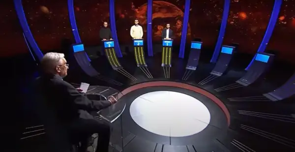 Trzech uczestników teleturnieju stoi przy swoich stanowiskach, a prowadzący siedzi przodem do nich.