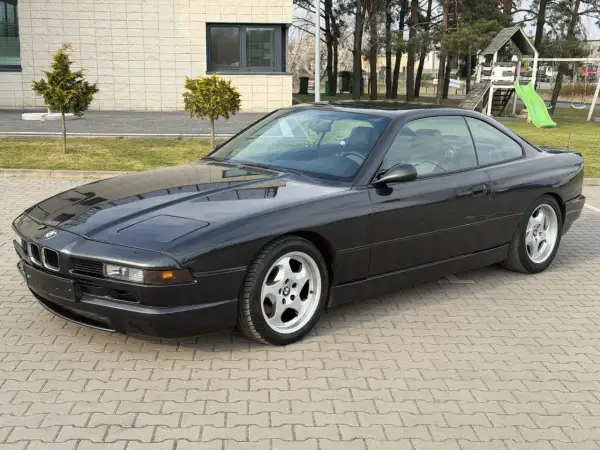 Czarne BMW V12, klasyk z lat 90., zaparkowane na kostce brukowej przed budynkiem z placem zabaw w tle.