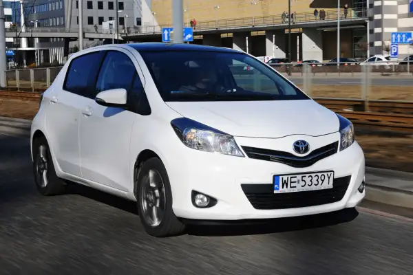 Białe małe auto, Toyota Yaris, jedzie po drodze. W tle budynki i torowisko tramwajowe.