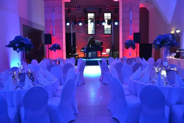 Elegancka filharmonia restauracja z fortepianem na scenie, stoły nakryte białymi obrusami i ozdobione kwiatami, oświetlone niebieskim i różowym światłem.