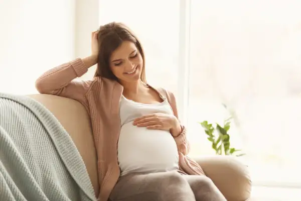 Uśmiechnięta ciężarna kobieta siedzi na kanapie, delikatnie dotykając brzucha. Myśli o bezpiecznym oczyszczaniu wodorowym w ciąży.