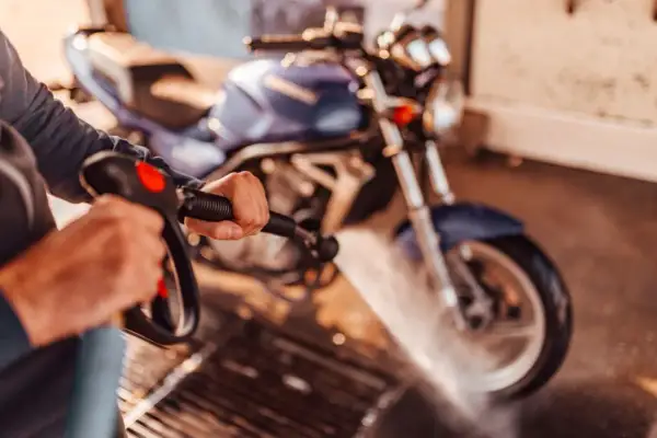 Persona limpiando una moto azul con una hidrolavadora. El agua salpica sobre la rueda delantera.