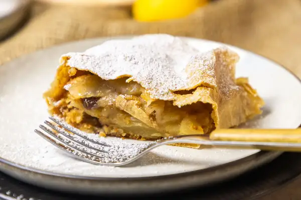 Ein Stück Apfelstrudel, bestäubt mit Puderzucker, wartet auf dem Teller. Dieses Apfelstrudel Rezept ist ein Genuss.