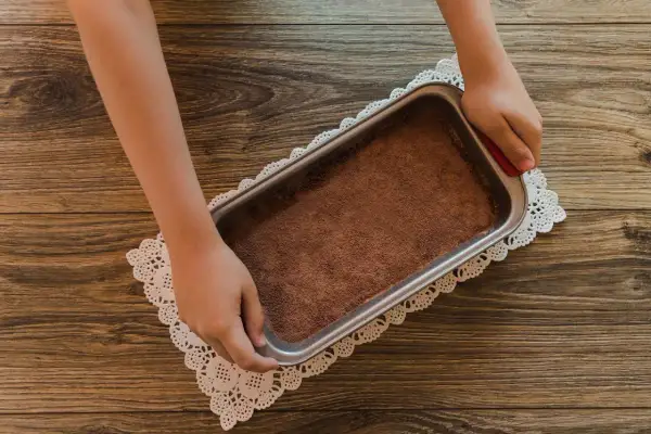Mani che preparano un tiramisù senza caffè, con cacao in polvere in una teglia su un centrino di pizzo.