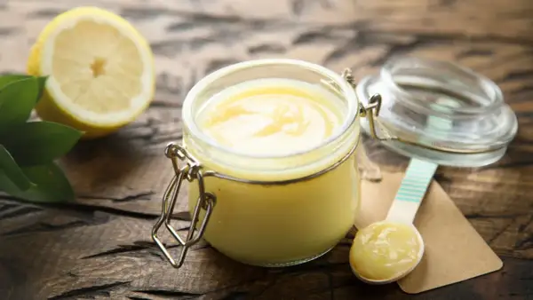 Un pot de lemon curd onctueux, une moitié de citron et des feuilles vertes sur fond de bois. Une cuillère en bois est prête à déguster.