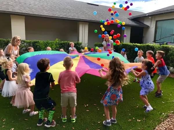 Children joyfully engage in gross motor activities for toddlers, tossing colorful balloons into the air while holding a large parachute.