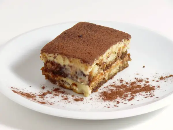 Une part de tiramisu, avec sa traduction française, est saupoudrée de cacao sur une assiette blanche.