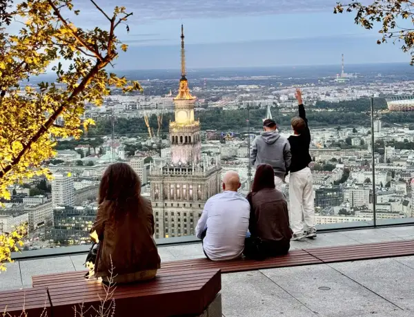 Ludzie podziwiają panoramę Warszawy z Pałacem Kultury i Nauki. Widać wiele warszawskich wież i budynków.