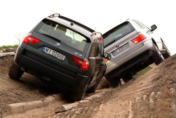Dwa BMW E83, zielone i srebrne, pokonują trudny teren, wspinając się po piaszczystym zboczu z drewnianymi przeszkodami.