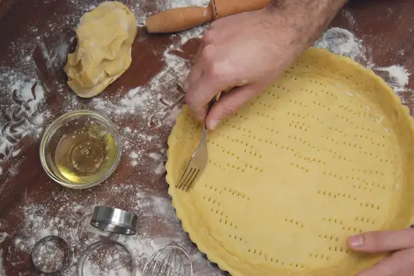 Mani che preparano una pasta frolla morbida, bucherellandola con una forchetta in una tortiera. Farina e ingredienti sparsi sul tavolo.