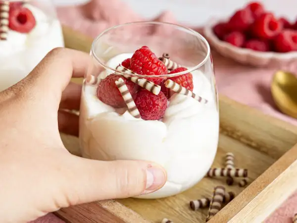 Ein Glas mit cremigem Quark-Dessert, garniert mit frischen Himbeeren und Schokoröllchen.