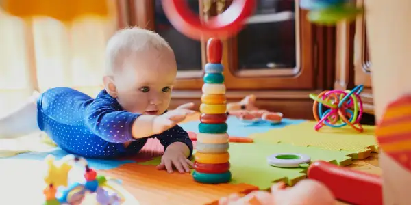 Zaintrygowany 6 miesięczne dziecko sięga do kolorowej wieży z kółek, otoczony zabawkami na macie.