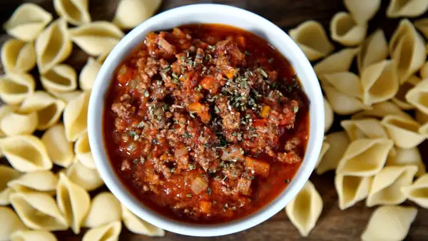 Herzhaftes Bolognese-Rezept mit Hackfleisch und Tomaten, garniert mit Kräutern, umgeben von ungekochten Nudelschalen.