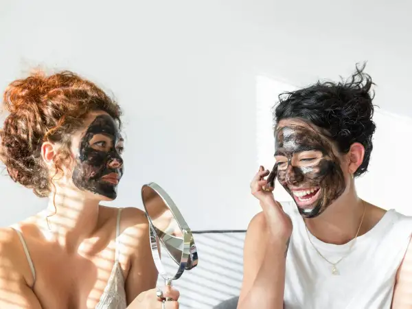 Dos mujeres con mascarilla negra en el rostro, una se mira en un espejo, buscando quitar espinillas negras grandes.