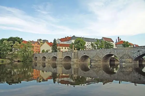 Historische Steinbrücke über den Fluss in Písek, Tschechien, mit Spiegelung im Wasser und bunten Häusern im Hintergrund.