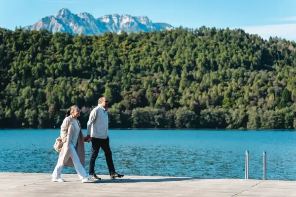 Coppia passeggia mano nella mano sul sentiero dei pescatori a Levico, con lago e montagne sullo sfondo.