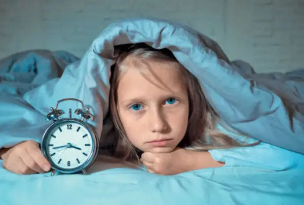 Smutna dziewczynka z niebieskimi oczami leży pod kołdrą obok budzika. Zmaga się z zaburzeniami snu.