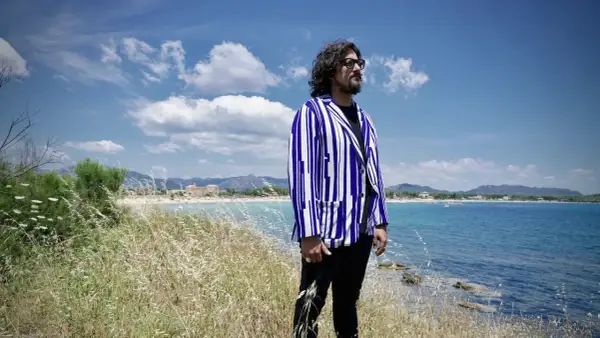 Uomo con giacca a righe blu e bianche guarda il mare in Sardegna, tra i campi di erba alta. Un'ambientazione perfetta per scoprire i **4 ristoranti Sardegna**.