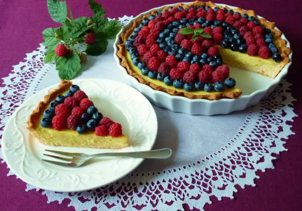 Pyszna tarta z kremem budyniowym, ozdobiona malinami i borówkami. Jeden kawałek już czeka na talerzyku.
