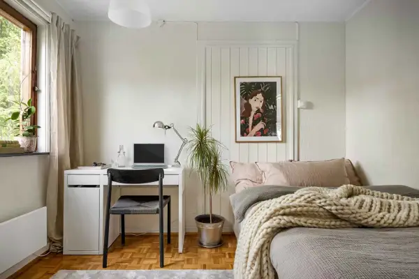 Gemütliches homeoffice im schlafzimmer mit Schreibtisch, Laptop und Bett. Ein Bild mit einer Frau und Pflanzen ziert die Wand.